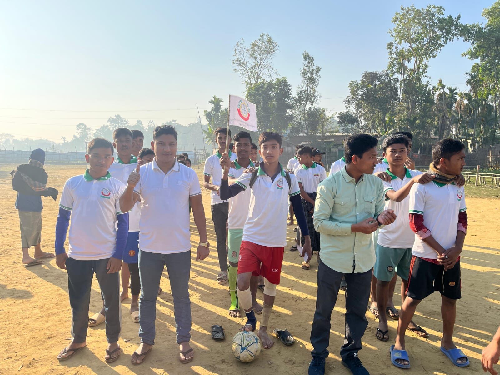 Players in school kit walking onto a dirt field behind the school flag in morning haze