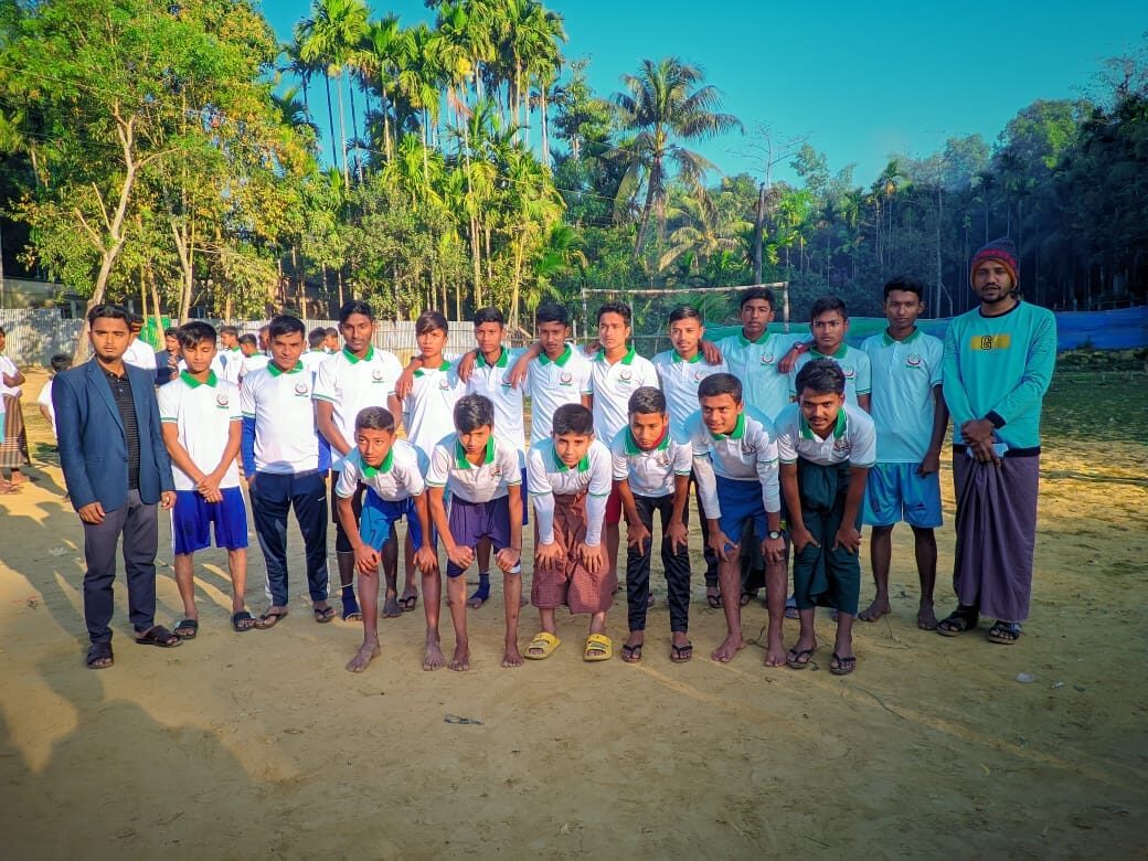 Team photo of students in school kit on the dirt pitch, palm trees behind